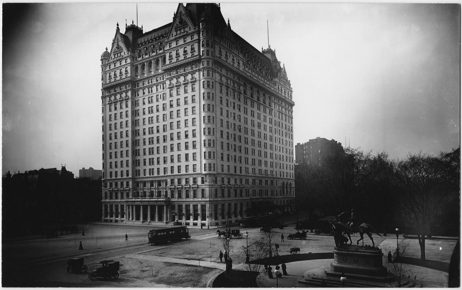 b&w photo of exterior of The Plaza Hotel with some people, horse carriages and cars